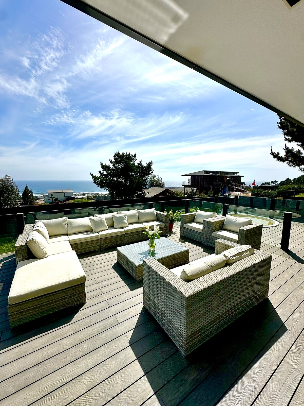 A spacious outdoor living area is shown, featuring comfortable seating arrangements with soft cushions. A coffee table is positioned at the center, surrounded by lush greenery and offering a view of the horizon and ocean. The deck is bathed in natural light.