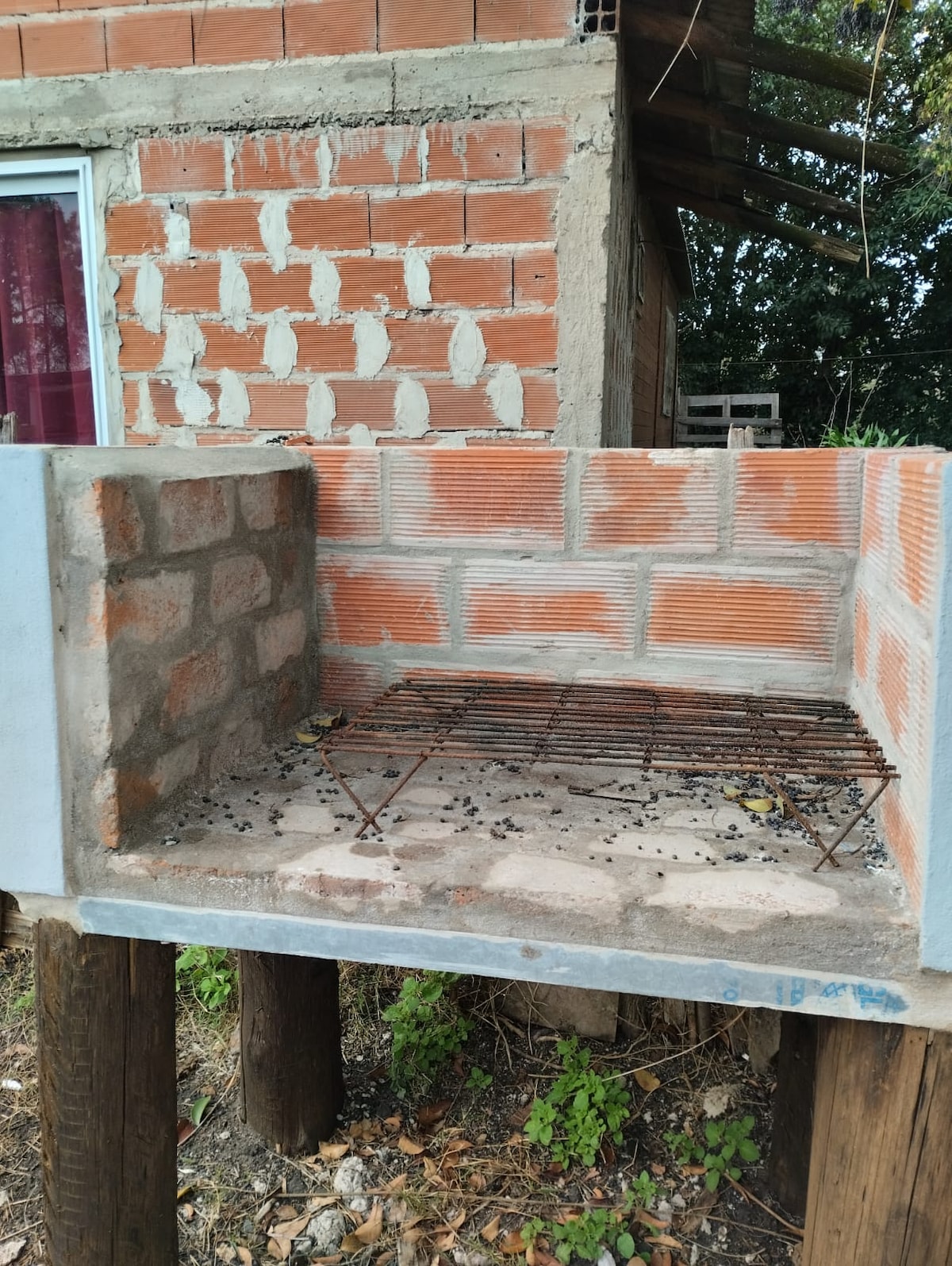 A brick barbecue grill is shown, featuring a stone base supported by wooden legs. The grill area is made with brick and includes a metal grate for cooking. Surrounding vegetation and a partially visible structure are also present in the background.