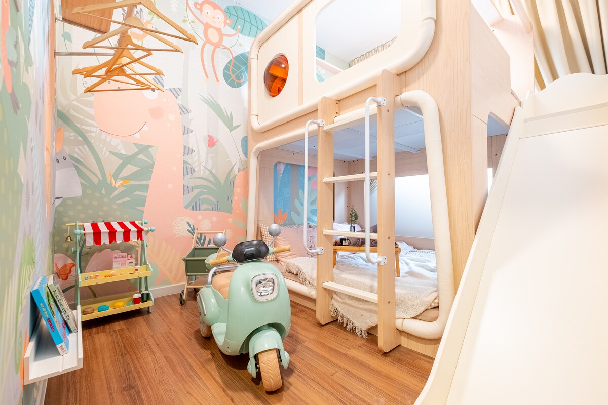 A playful children's bedroom features a double-decker bed with slides and a vibrant jungle-themed wallpaper. A toy scooter is positioned beside the bed, and a small play cart is visible, providing a cheerful atmosphere for play and rest.