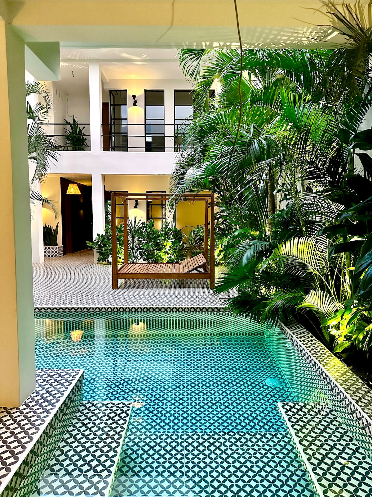 A vibrant courtyard is surrounded by lush greenery, showcasing an elegant pool with intricate black and white tile patterns. A wooden swing can be seen nearby, inviting relaxation amidst the tropical atmosphere. Natural light brightens the space, emphasizing the tranquil setting.