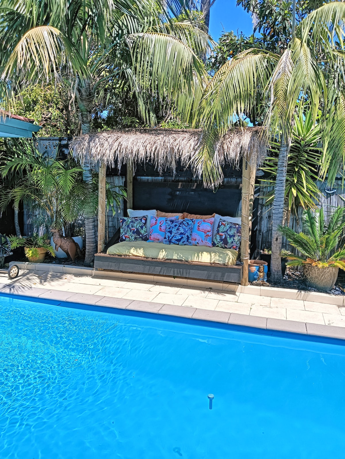A shaded Bali daybed is positioned beside a sparkling blue swimming pool, surrounded by lush greenery and tropical plants. Decorative cushions adorn the daybed, providing a comfortable space to relax in the outdoors.