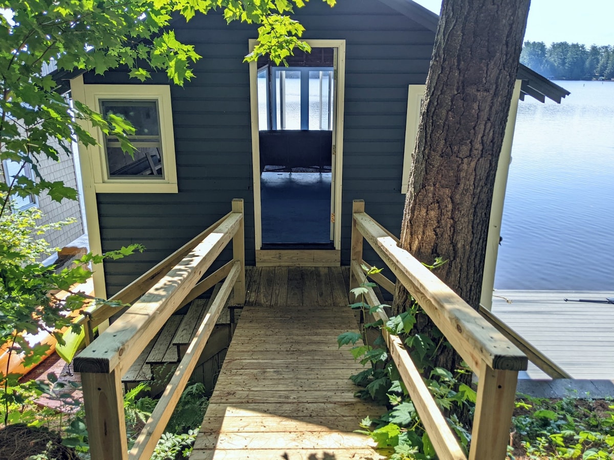 A wooden walkway leads from a grassy area to the entrance of a boat house. The house is surrounded by trees and offers views of the lake from large windows. Light streams through the open door, inviting guests inside.
