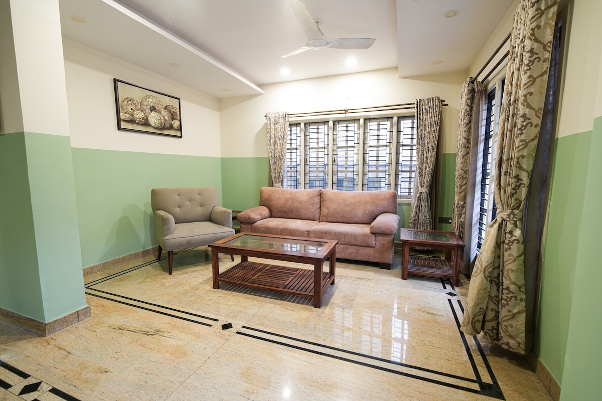 A living area features a plush sofa and a comfortable armchair, arranged around a wooden coffee table. Natural light filters through the large windows, adorned with patterned curtains. The marble floor is complemented by a clean, neutral color scheme on the walls.