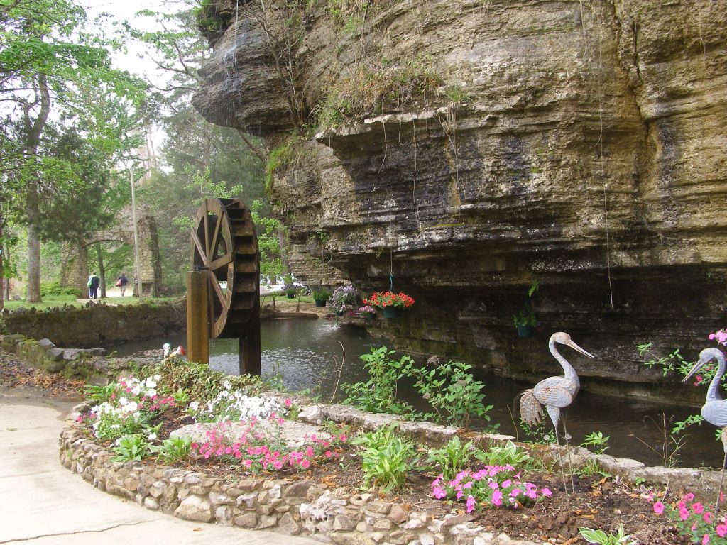 A water wheel is positioned near a tranquil pond, surrounded by lush greenery and colorful flowers. Stone-lined flower beds enhance the natural beauty, while decorative cranes add a charming touch to the serene landscape.