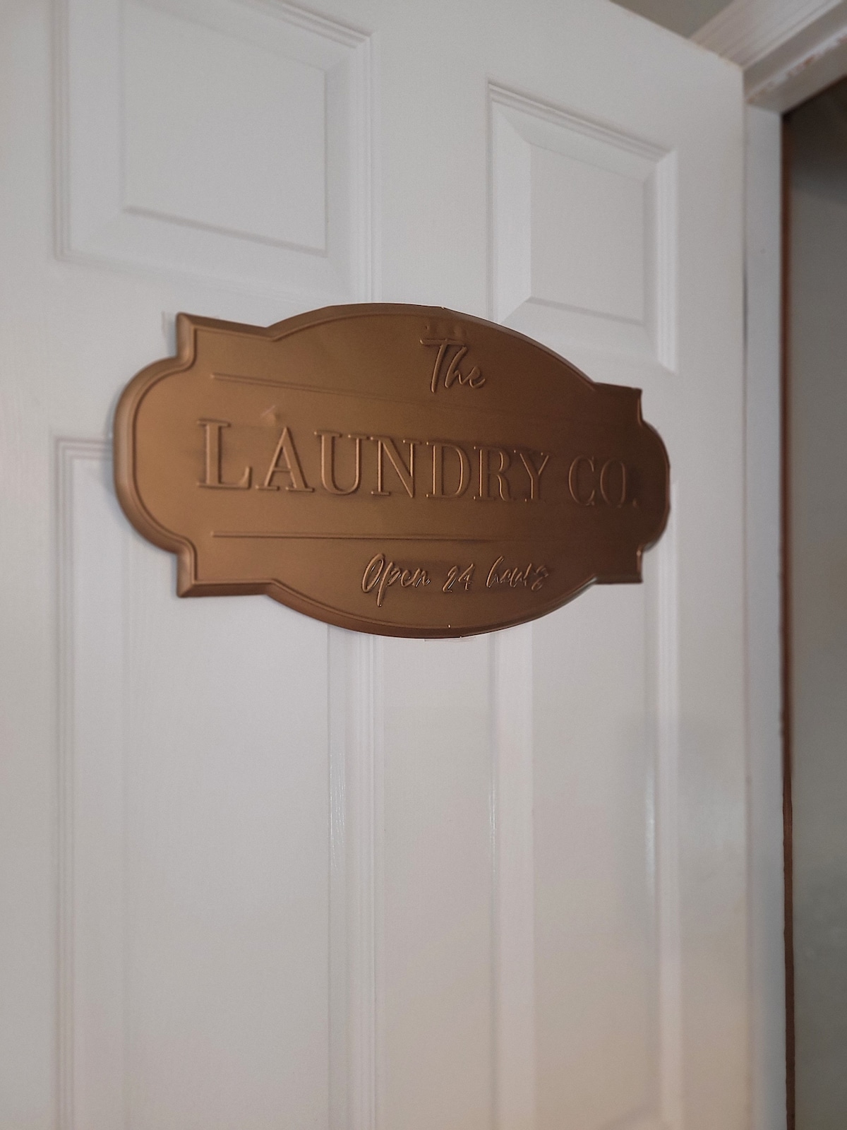 A bronze sign, reading 'The Laundry Co. Open 24 Hours', is mounted on a white door. The door features a classic panel design, complementing the sign's elegant typography.