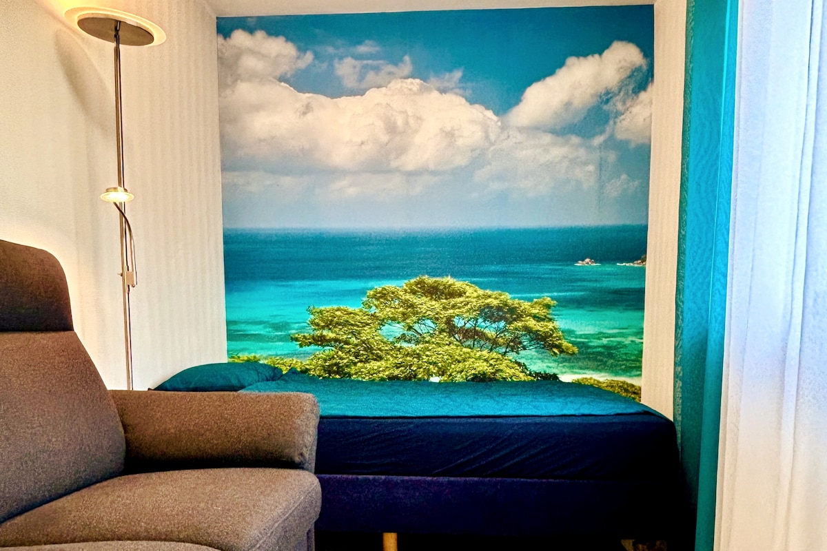 A serene mural depicting a blue ocean and lush tree is featured on one wall, enhancing the calming ambiance. A spacious bed is situated next to a comfortable chair under a soft lighting fixture, allowing for relaxation and leisure.