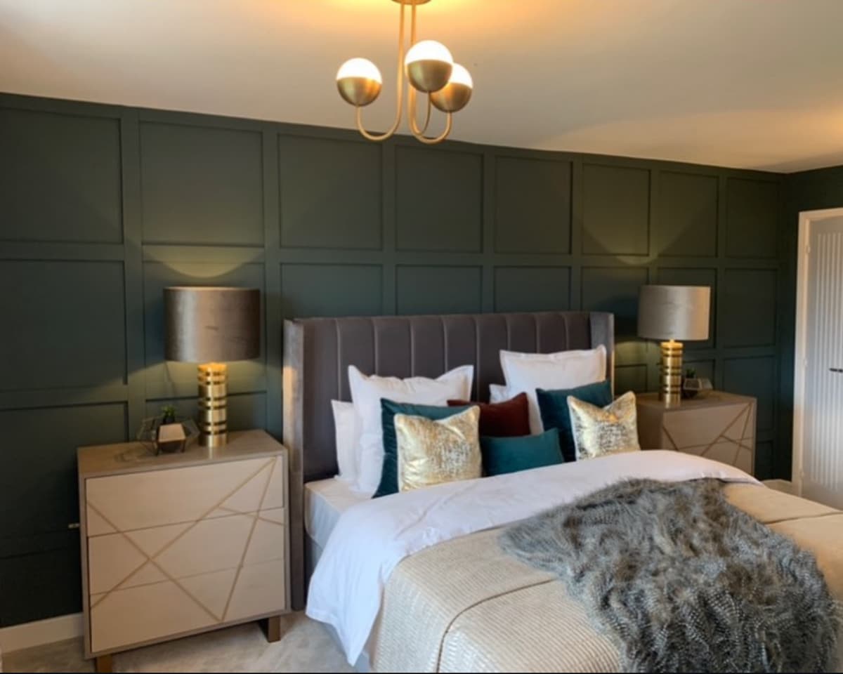 A stylish bedroom features a plush bed with layered pillows in varying textures. Two modern lamps flank the bed on sleek nightstands. The dark green accent wall and cozy throw add depth and warmth to the inviting space.