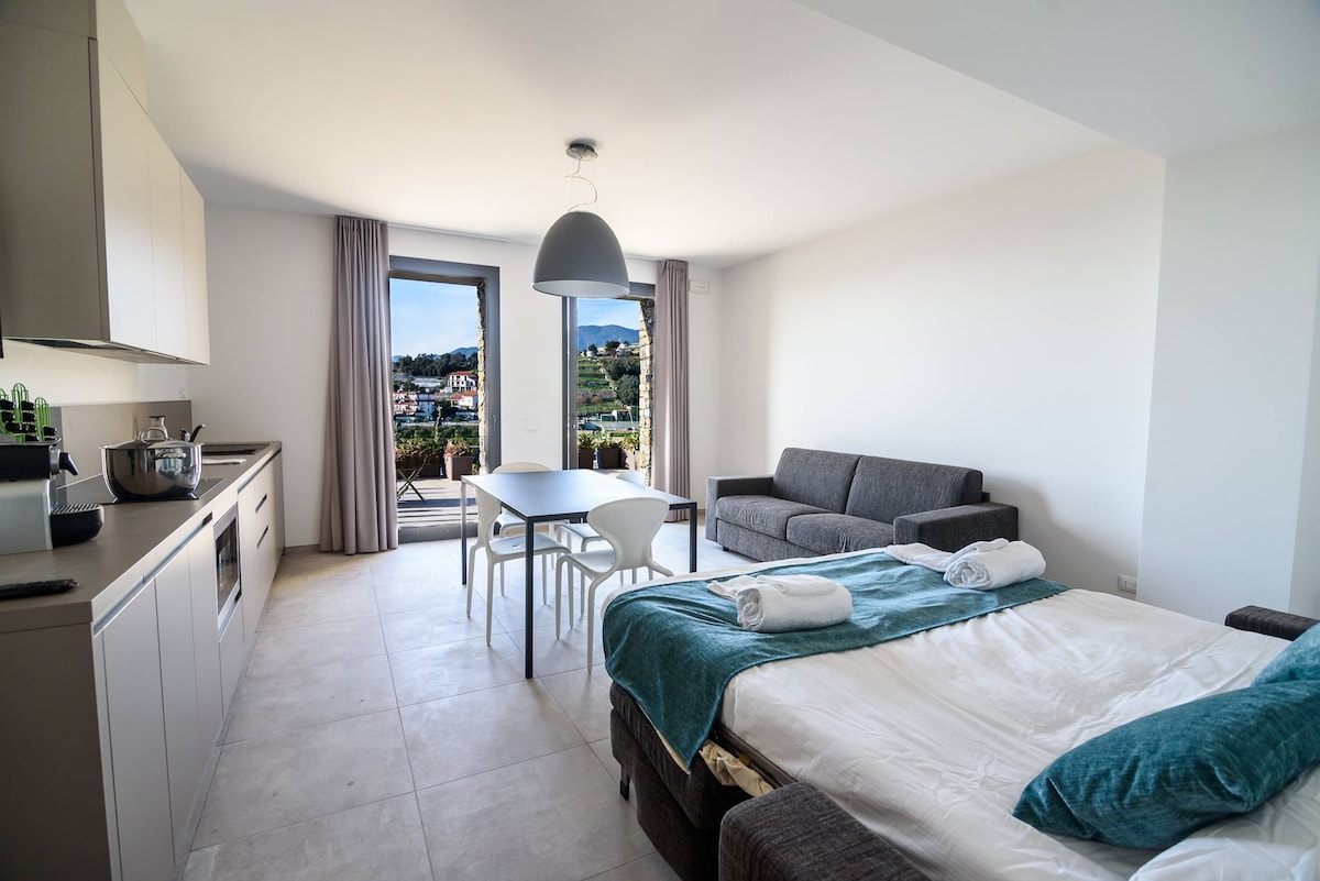 A bright living area is presented, featuring a spacious bed with a turquoise throw and neatly arranged towels. A dining table with four chairs is nearby, complemented by a modern kitchen. Large windows provide views of the landscape outside, enhancing the sense of openness.