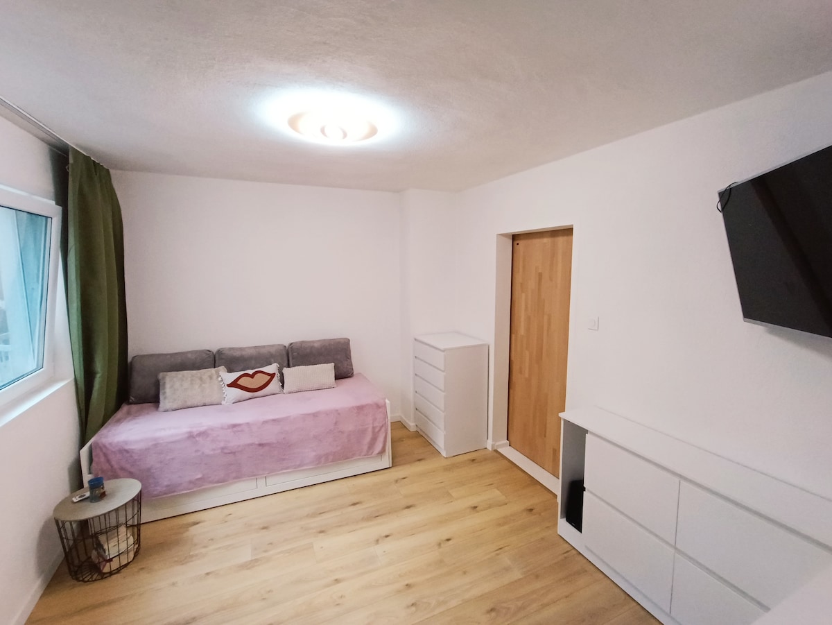 A light-filled living space features a spacious sofa with a pink cover and decorative cushions. A minimalist white dresser and TV mount add functionality. Natural light enters through a nearby window, complemented by a green curtain. A small round table enhances the cozy atmosphere.