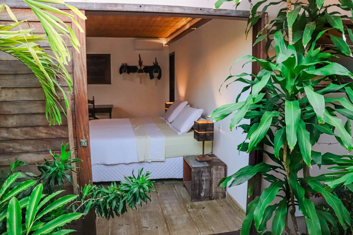 A serene bedroom is framed by lush greenery, featuring a spacious bed with light-colored linens. Soft lighting is provided by bedside lamps on rustic wooden nightstands. The natural wood flooring adds warmth, and the decorated walls hint at a calming, nature-inspired theme.