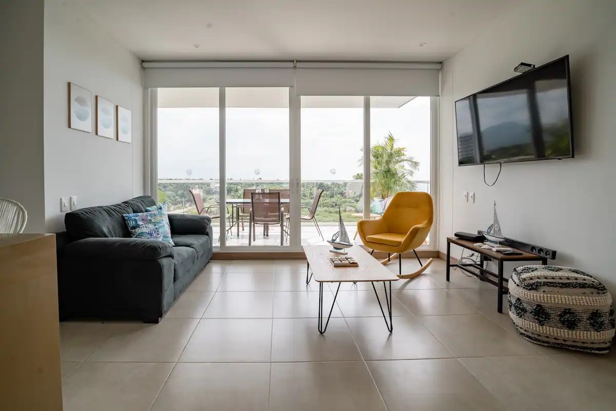 A living area features a dark sofa and a bright yellow armchair, with a coffee table positioned between them. Large sliding glass doors open to a balcony with outdoor seating, providing views of greenery and distant hills. A television is mounted on the wall.