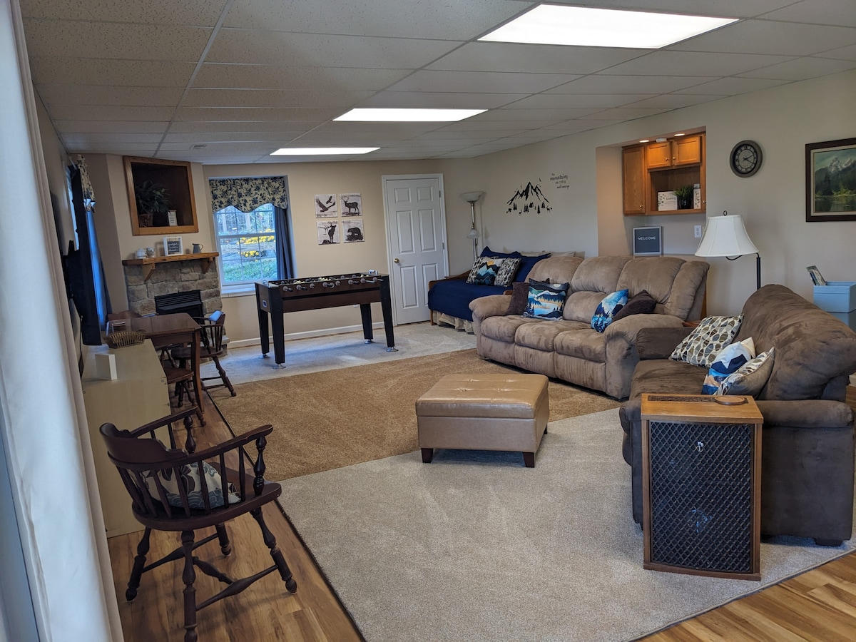 A spacious living room features a large sectional sofa and a coffee table at the center. A foosball table is visible nearby. Light streams in from windows facing the yard, complemented by a fireplace and some wall decor. Comfortable chairs add to the inviting space.