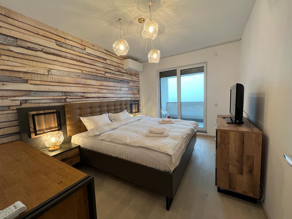 A spacious bedroom features a large bed framed by a textured wooden wall. Illuminated by stylish pendant lights, the room includes a dresser and a television. Glass doors provide access to a balcony, allowing natural light to enhance the serene ambiance.