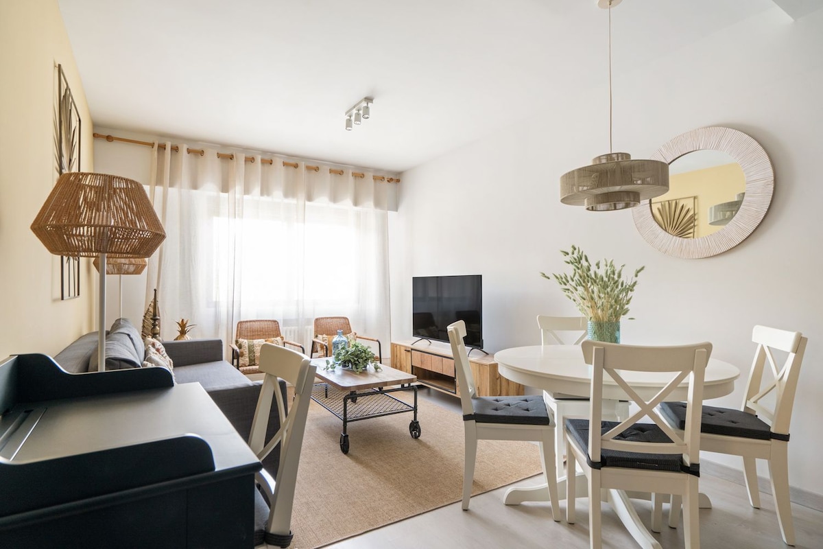 The living area features a combination of a comfortable sofa and two accent chairs, arranged around a central coffee table. A round dining table sits adjacent to a large window, allowing natural light to brighten the space. A wall-mounted TV and decorative elements add character.