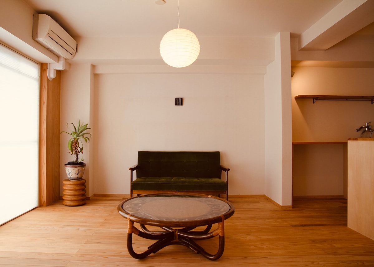 The living area features a green sofa positioned in front of a large window, allowing natural light to fill the space. A circular coffee table with a woven design stands at the center, and a potted plant adds a touch of greenery. A simple pendant light hangs above.