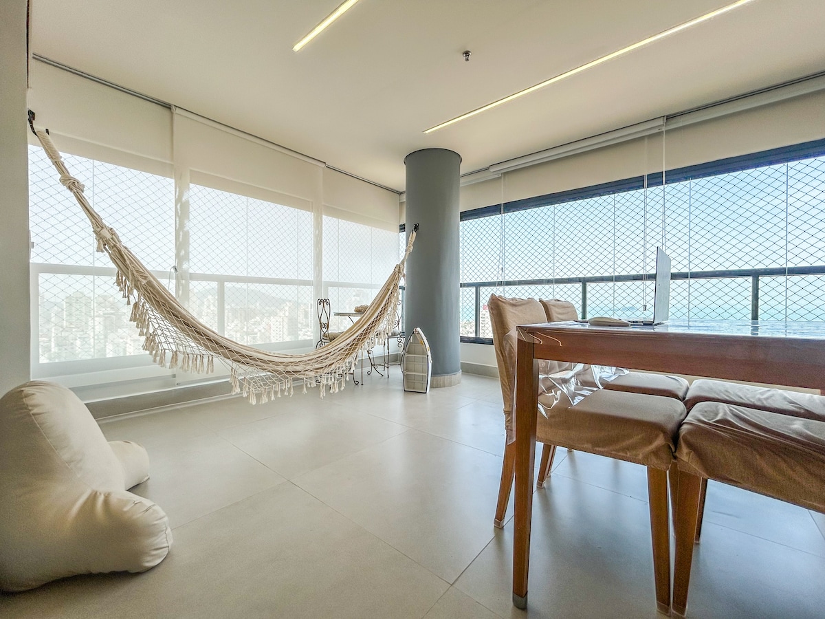 A comfortable living area features a hammock suspended between two supports, providing a cozy relaxation space. A wooden table and chairs are positioned nearby, along with a soft bean bag. Large windows offer panoramic views, complemented by natural light filtering through the window coverings.