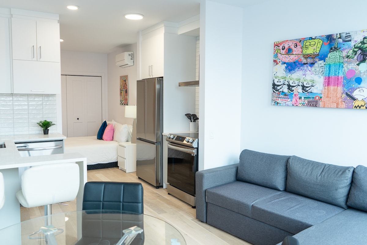An open-plan living space includes a modern kitchen with stainless steel appliances. A cozy sofa facing a colorful wall art piece complements a comfortable sleeping area in the background. A round glass dining table is positioned with black chairs, enhancing the contemporary design.