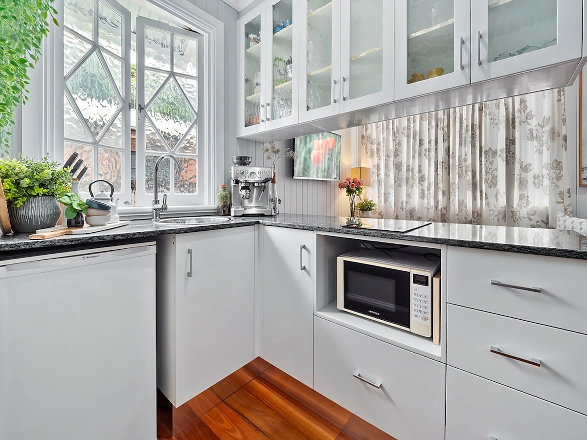 A well-equipped kitchen features modern cabinetry and a sleek countertop. A compact microwave is integrated into the cabinetry, while a coffee machine and sink are easily accessible. Natural light filters through a large window, complemented by decorative plants and soft window treatments.
