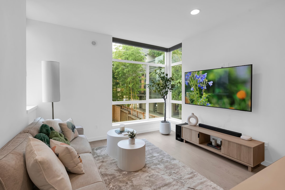 A bright living room features a light-colored sofa adorned with patterned cushions. A large flat-screen TV is mounted on a light wooden media console. Natural light fills the space through expansive windows overlooking greenery, complemented by a minimalistic floor lamp and circular coffee tables.
