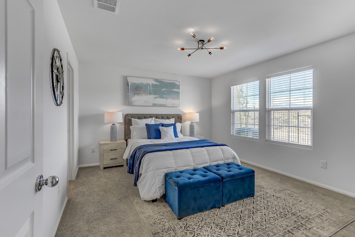 A well-lit bedroom features a spacious bed dressed in white linens with a blue throw. Two nightstands with lamps flank the bed, and large windows allow natural light to fill the room. A decorative area rug lies beneath, adding a touch of warmth.