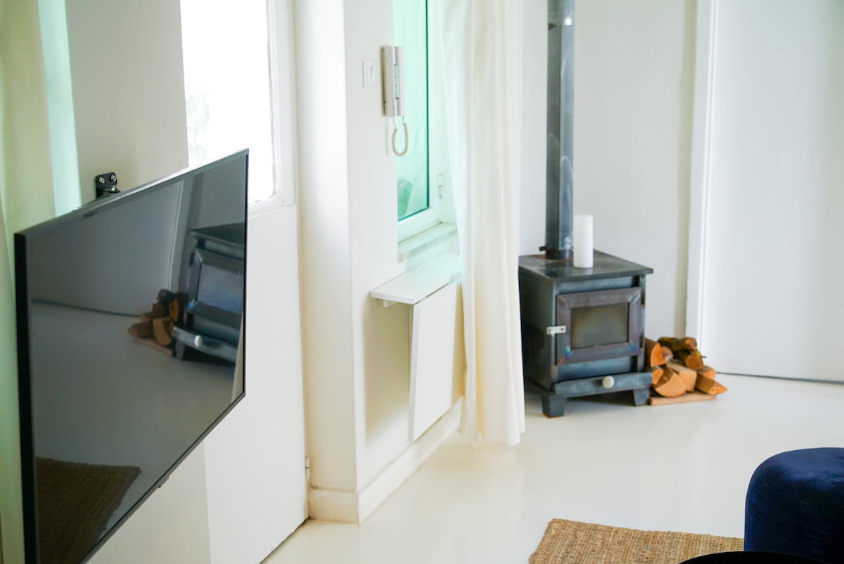 A living space is visible featuring a flat-screen television mounted on a white wall. A wood-burning stove sits nearby on a white surface, accompanied by neatly stacked firewood. Natural light filters through a window framed by sheer curtains.