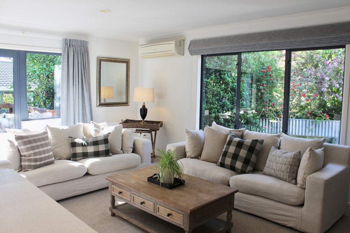 A bright living area is presented with comfortable, light-colored sofas adorned with various patterned cushions. A wooden coffee table occupies the center, while natural light enters through expansive glass doors, revealing greenery outside. A decorative lamp and mirror add to the inviting ambiance.