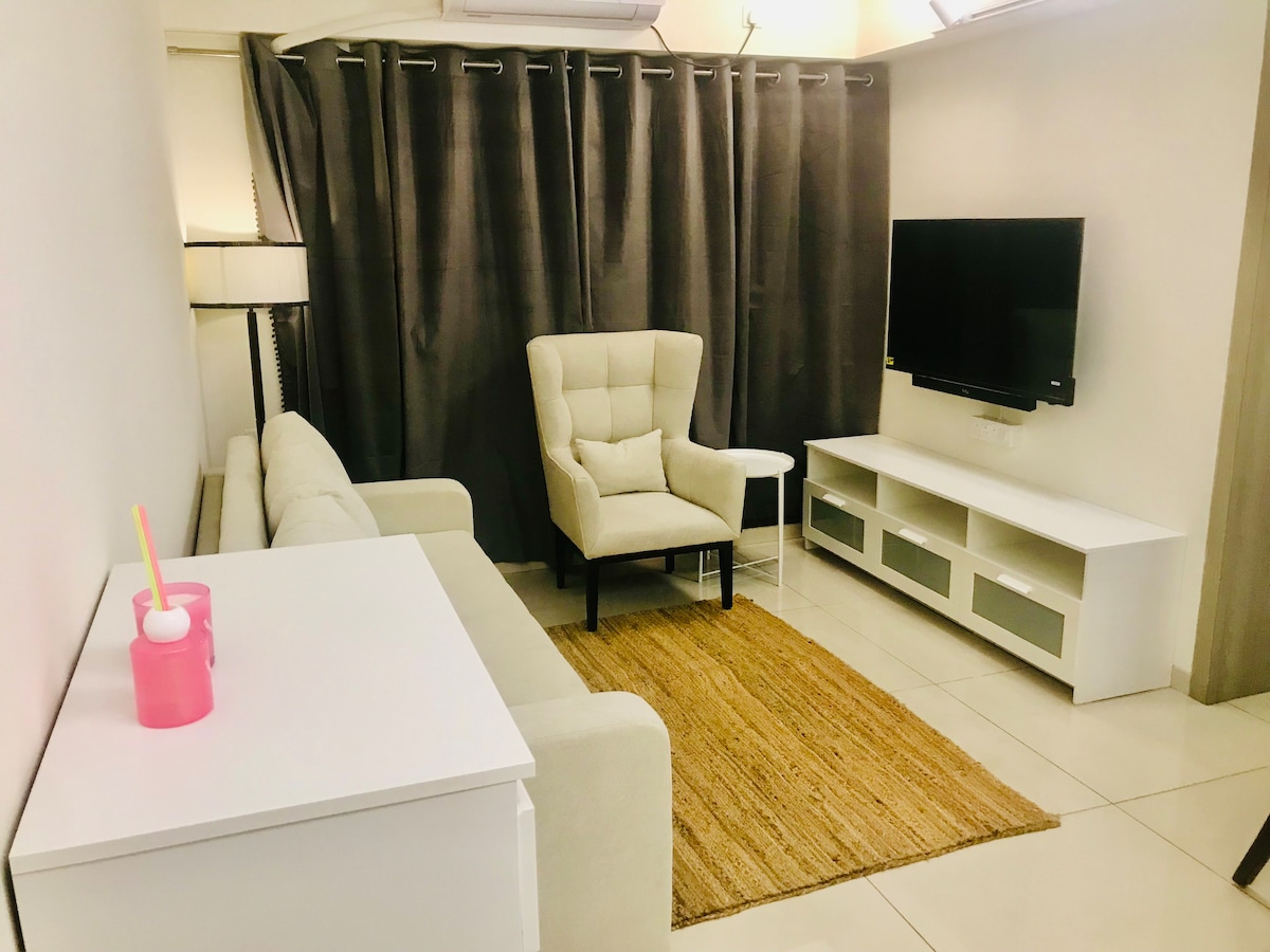 A spacious living area features a light-colored sofa and a comfortable armchair positioned near a wall-mounted television. A coffee table sits beside the sofa, accompanied by a textured area rug. Curtains in a dark shade frame the window, enhancing the cozy atmosphere.