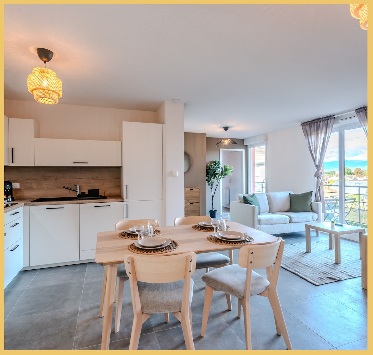A modern living space is showcased, featuring a dining area with a light wooden table set for four. The adjacent kitchen, equipped with essential appliances, complements the open floor plan. Natural light floods in from the balcony, enhancing the cozy seating area with a comfortable sofa.