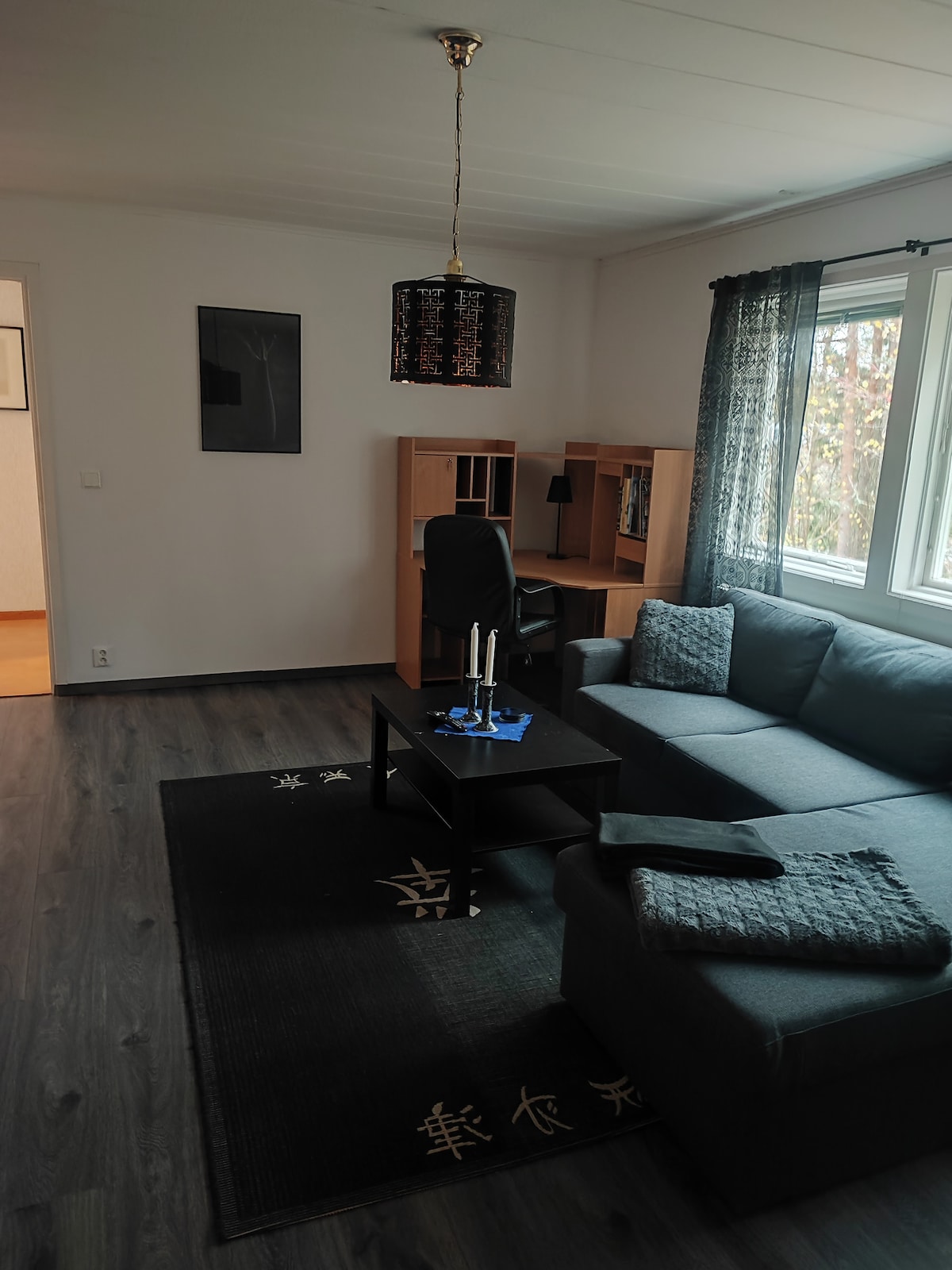 A living area is presented with a dark gray sofa positioned alongside a small black coffee table. A chair and shelving unit are positioned nearby, and a candle holder with candles adorns the table. Natural light is filtered through the large window, draped with sheer curtains.