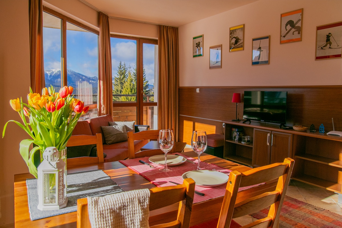 A bright dining area features a wooden table elegantly set with plates and glasses. A vase of tulips adds a vibrant touch. The room is bathed in natural light from large windows offering views of the mountains. A cozy lounge area is also visible.