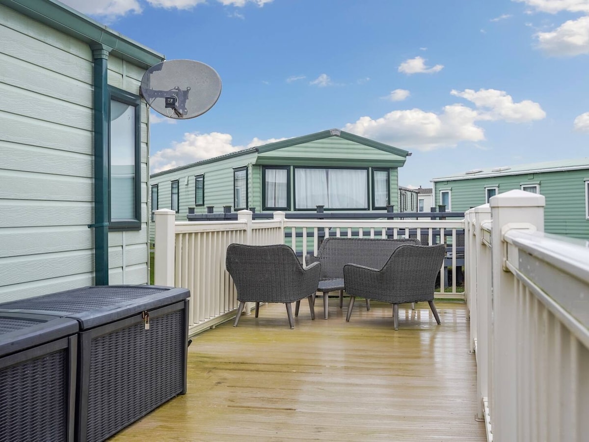 An enclosed decking area is visible, featuring two rattan-style chairs facing outward. A small table is positioned between them. A storage unit is also present, and the surrounding area showcases other caravans and a clear blue sky.