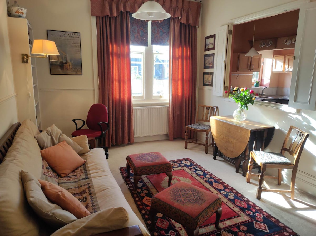The living area features a comfortable sofa adorned with decorative cushions, complemented by two wooden chairs and a small round table. Large windows are draped with red curtains, allowing natural light to illuminate the space. A colorful rug adds warmth to the floor.