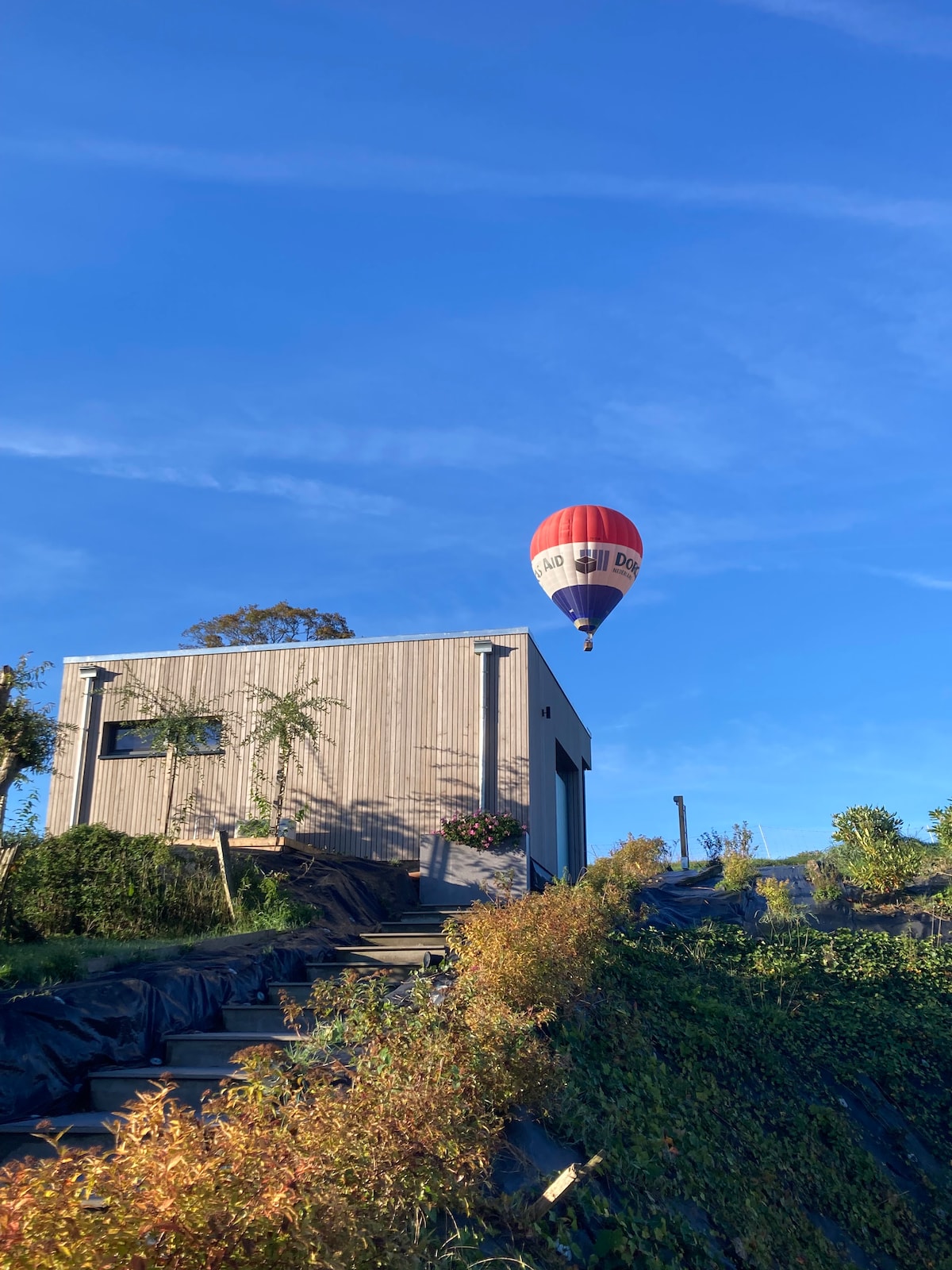 A modern wooden house sits on a hillside, surrounded by greenery. Above, a vibrant red and white hot air balloon drifts in the clear blue sky. Steps lead from the house down the slope, which is dotted with plants.