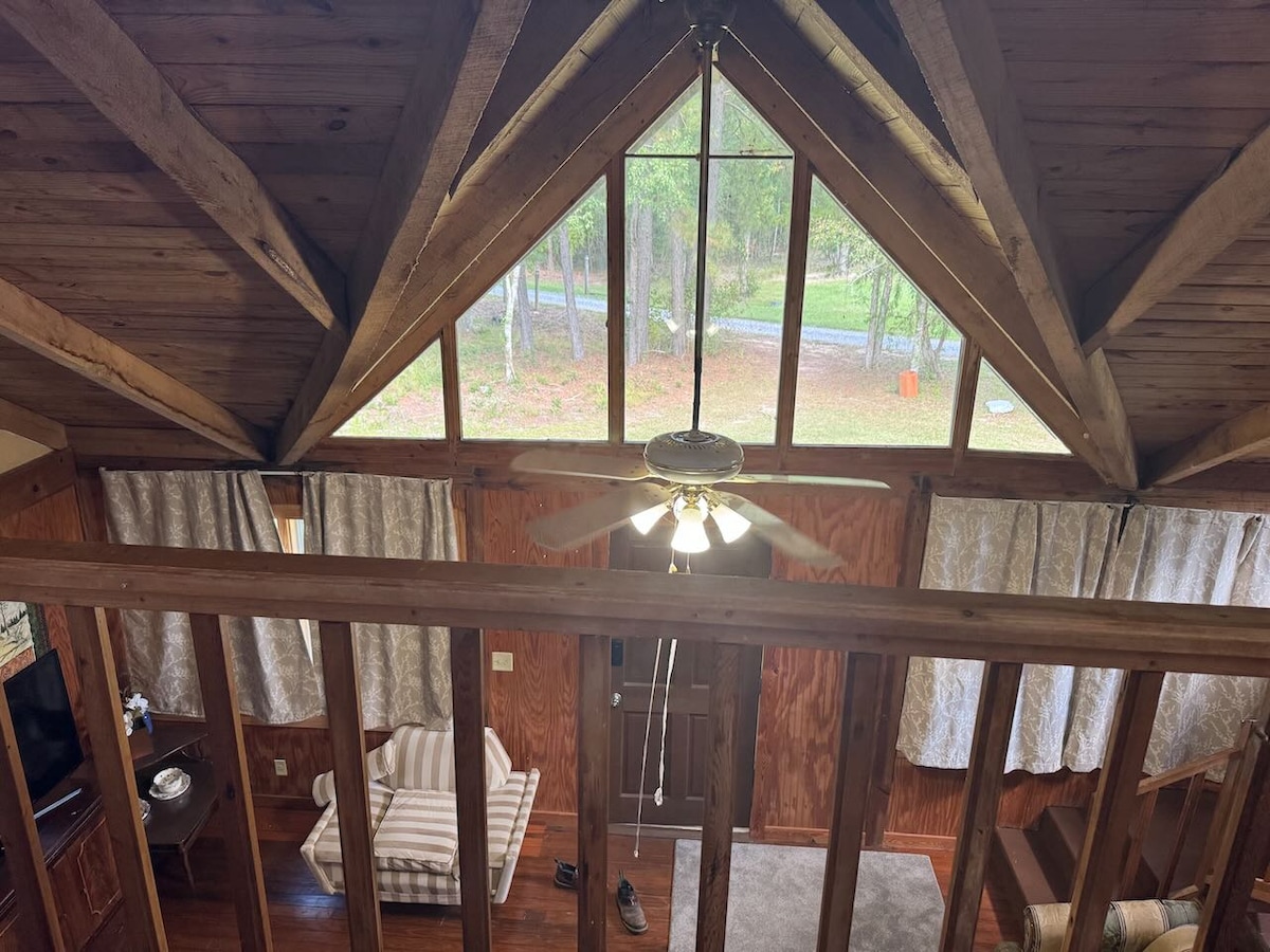 The interior of the cabin features a vaulted ceiling with exposed wooden beams. Large windows allow natural light to fill the space, revealing views of the surrounding trees. A ceiling fan hangs above, and cozy seating is visible on the lower level.