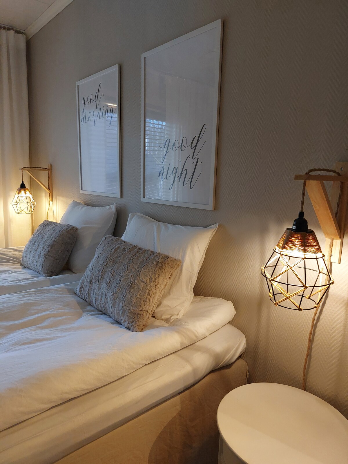 A serene bedroom features two spacious beds with light bedding, adorned with decorative pillows. Soft lighting from unique bedside lamps creates a calming atmosphere. Two framed wall prints displaying the phrases 'good morning' and 'good night' complement the design.