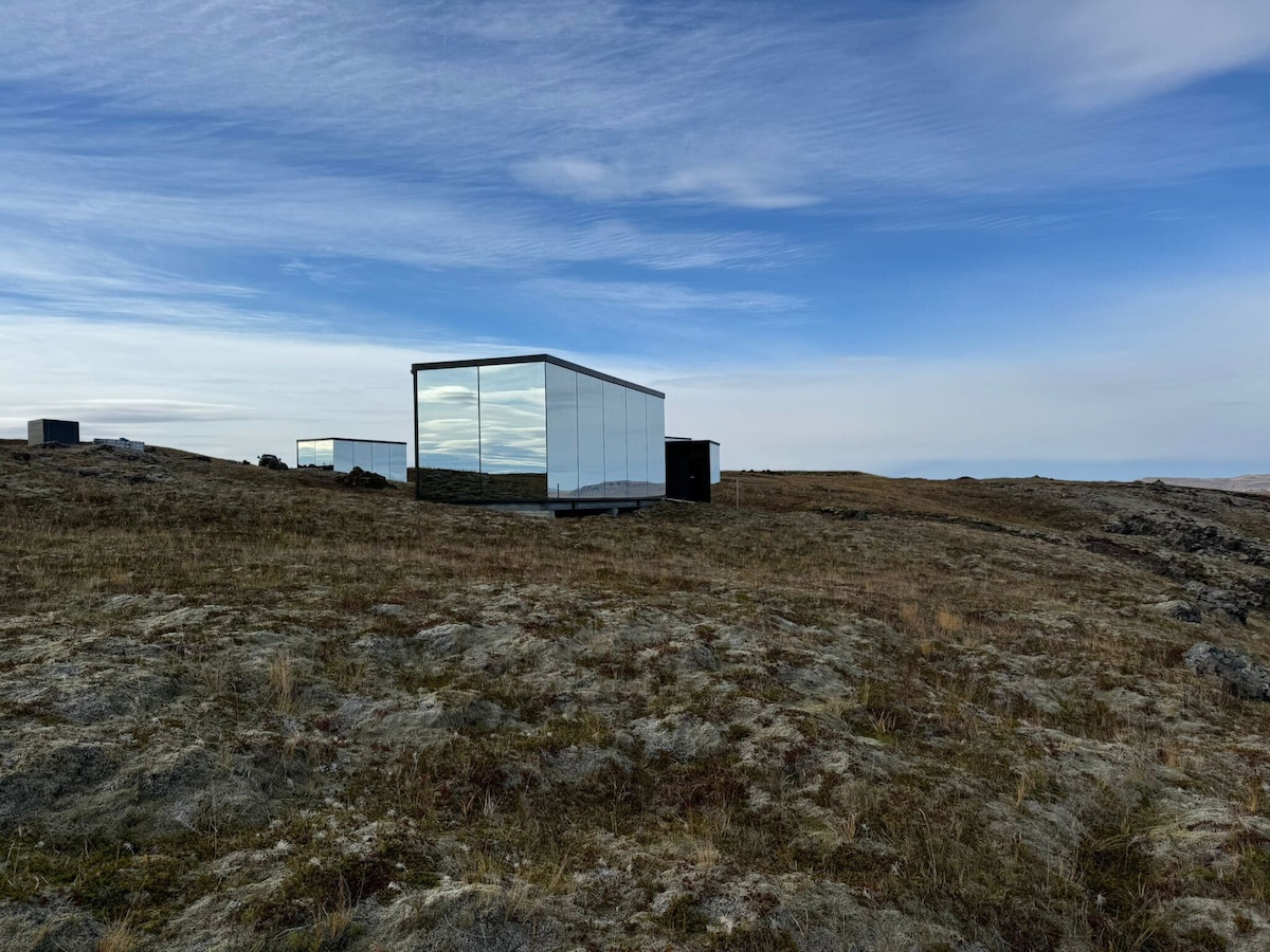 A modern glass structure is depicted on a grassy terrain, reflecting the surrounding landscape. The building's sleek design contrasts with the rugged moss-covered ground under a clear blue sky, showcasing the harmony between architecture and nature.