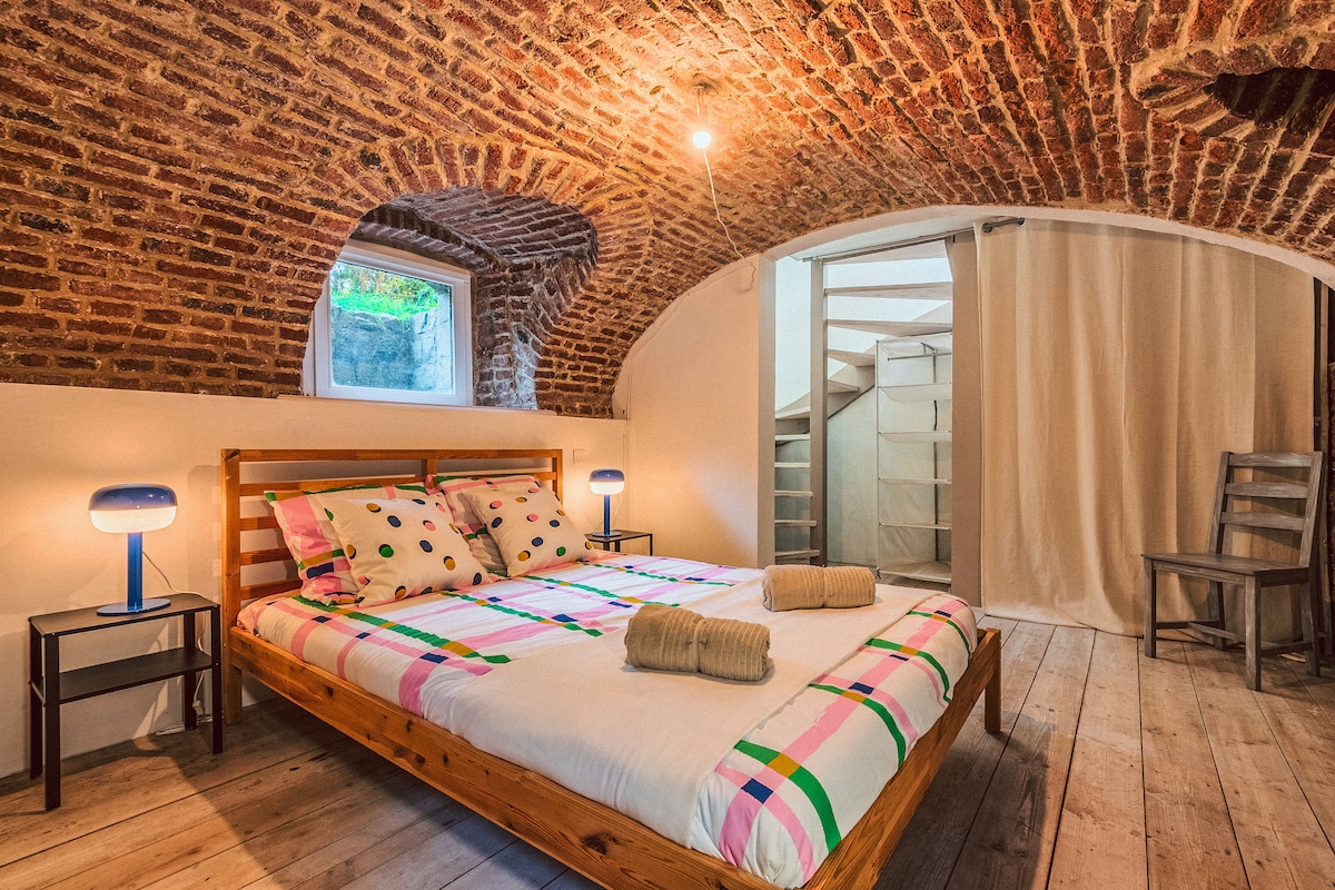 An inviting sleeping area features a spacious bed adorned with colorful patterned linens. Exposed brick arches create an atmospheric backdrop. A window offers natural light, and a simple wooden chair and side tables with lamps complete the cozy ambiance.