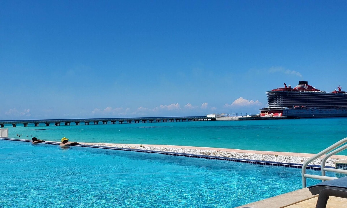A large infinity pool extends towards a clear blue ocean, reflecting the vibrant sky. A cruise ship is docked in the distance, with a wooden pier leading into the water. Sun loungers are positioned nearby for relaxation.