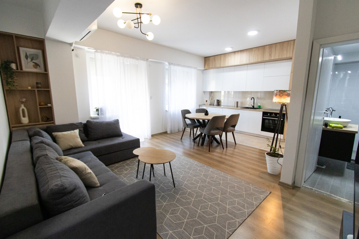 A stylish living area features a large gray sofa, complemented by a circular wooden coffee table in front. A dining table with chairs is positioned nearby, showcasing a modern kitchen equipped with sleek cabinetry. Natural light is filtered through sheer curtains, illuminating the space.