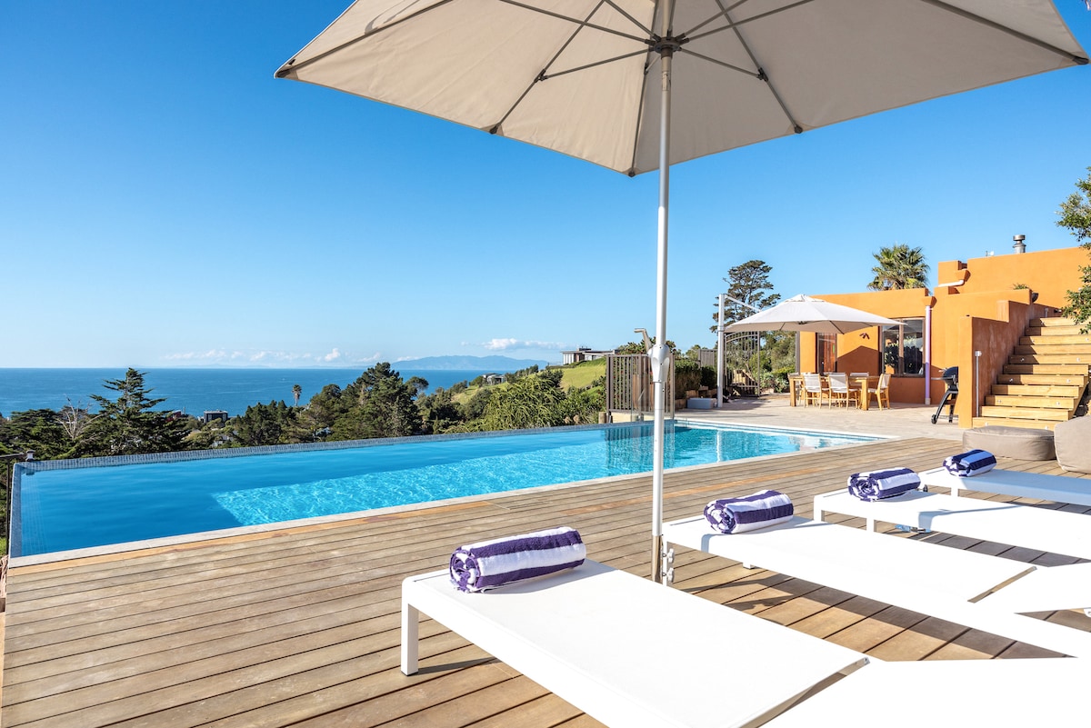 A serene outdoor area features a private swimming pool surrounded by wooden decking. Sun loungers with rolled towels are positioned under large umbrellas, providing shade. The ocean view stretches into the distance, complementing the clear blue sky and well-maintained gardens.