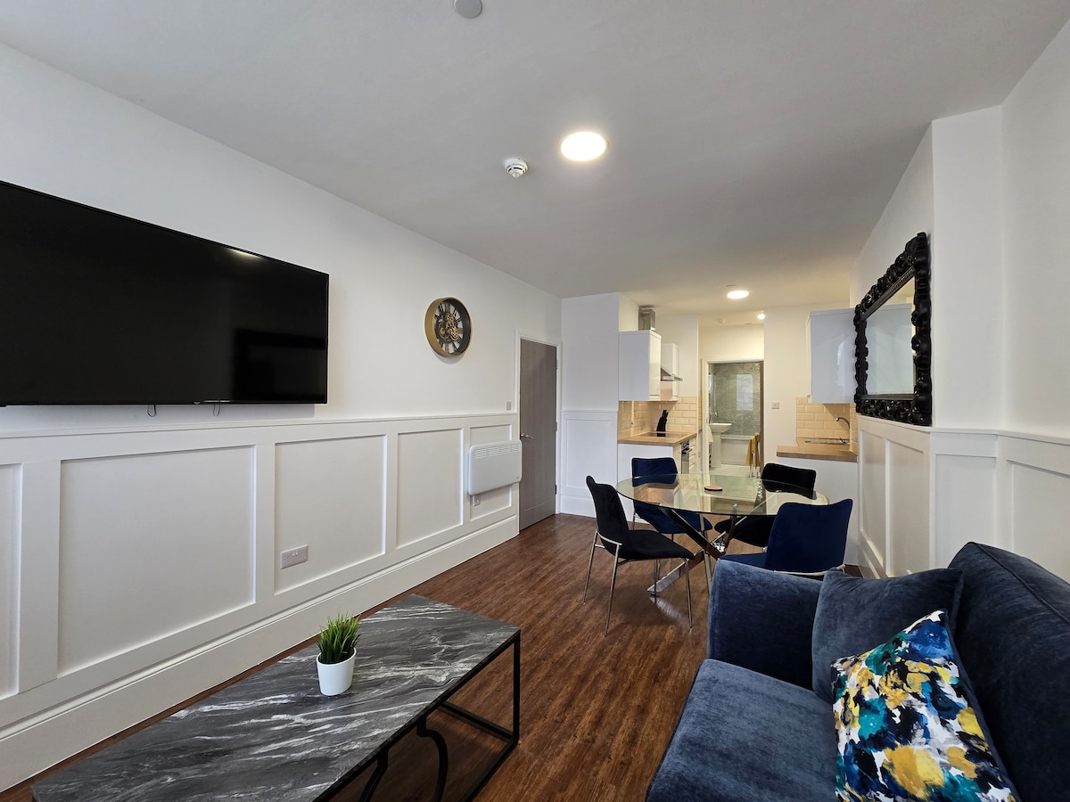The living area features contemporary furnishings, including a comfortable sofa and stylish chairs surrounding a glass table. A large television is mounted on the wall, while decorative elements and a wall clock add subtle character to the space. Natural light enhances the warmth of the room.