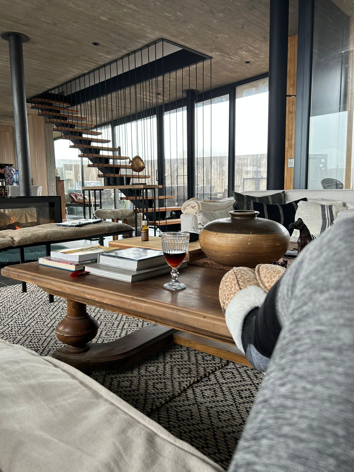 A modern living area features a wooden coffee table set with books and a decorative bowl. A glass of red beverage rests on the table. A comfortable seating arrangement with neutral tones surrounds a cozy fire, and a spiral staircase is visible in the background.