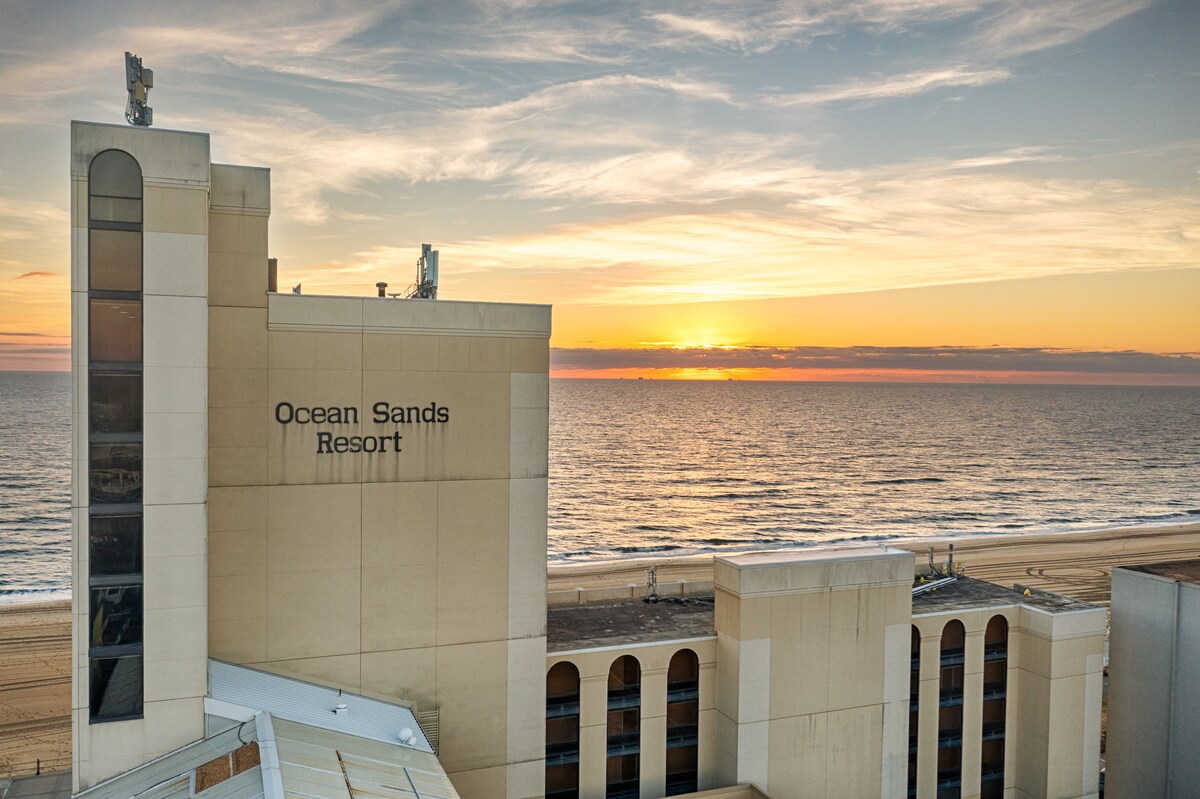 The exterior of Ocean Sands Resort is showcased against a sunset backdrop, with the sun reflecting off the water. The building features a modern design with large windows, and the resort name is clearly displayed.