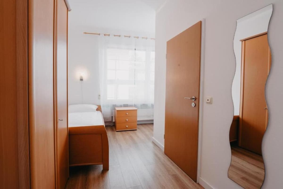 A single room is presented, featuring light wood furnishings and a spacious bed. A wardrobe is positioned near the entrance, accompanied by a bedside table. Natural light is allowed in through a large window with sheer curtains. A full-length mirror enhances the room's sense of space.