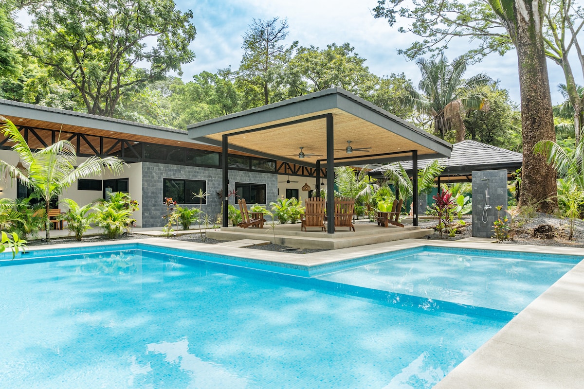 A spacious villa is surrounded by tropical greenery, featuring a large swimming pool with clear water. The outdoor area includes a covered lounge space with wooden seating. The architecture shows a blend of open spaces and natural surroundings, offering a tranquil vacation setting.