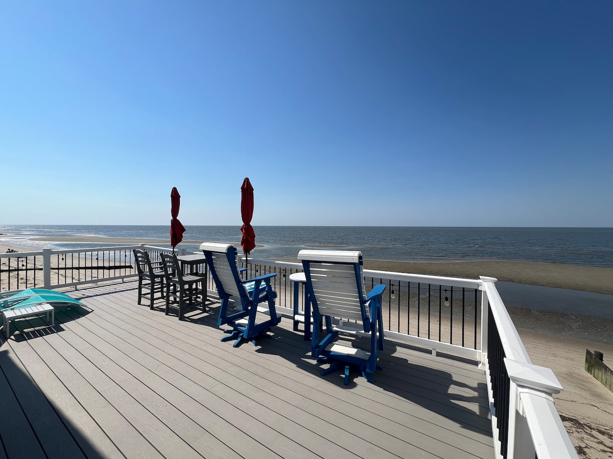A sunlit deck overlooks the shoreline, featuring a row of patio chairs in blue and dark wood. Two red umbrellas provide shade, enhancing the seating area. The calm sea and clear blue sky stretch into the horizon, creating a serene coastal backdrop.