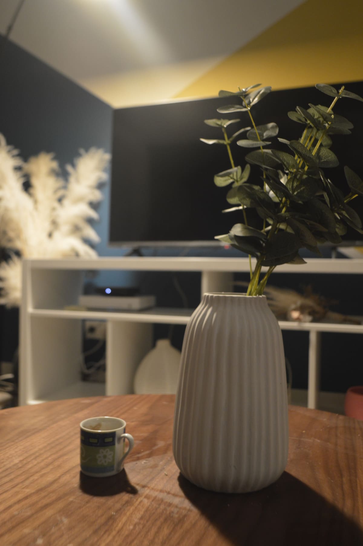 A wooden table features a textured white vase filled with greenery, positioned in front of a television on a low, modern shelf. A small decorative cup sits beside the vase, adding a subtle touch to the scene, which is illuminated by soft lighting.