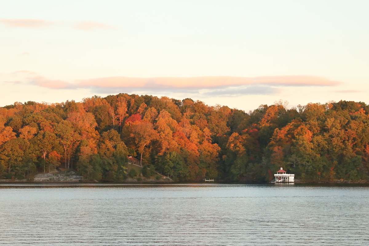 A serene waterfront view captures a variety of autumn-colored trees, reflecting warm hues across the water. A gazebo is positioned on the shore, contrasting with the vibrant foliage that lines the landscape, under a softly lit sky.