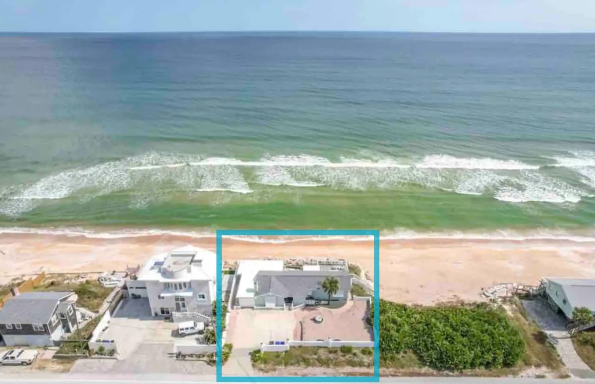 An aerial view captures the Seaglass Cottage nestled along the shore, featuring a spacious patio area outlined by light-colored pathways. The ocean's gentle waves are visible, accentuating the home's prime beachfront location, with neighboring houses positioned in a tranquil seaside setting.