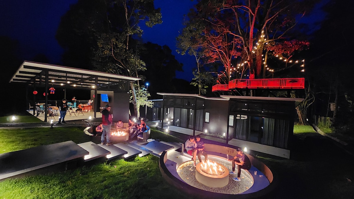 An outdoor gathering is depicted, featuring multiple guests seated around a circular firepit. The scene is illuminated by soft lighting from overhead string lights and the glow of the fire. Nearby, modern cabins with large windows sit surrounded by lush greenery, offering a serene backdrop.